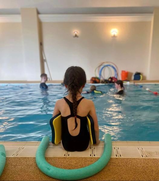 A girl sitting beside the pool, looking towards a group lesson taking place in the water