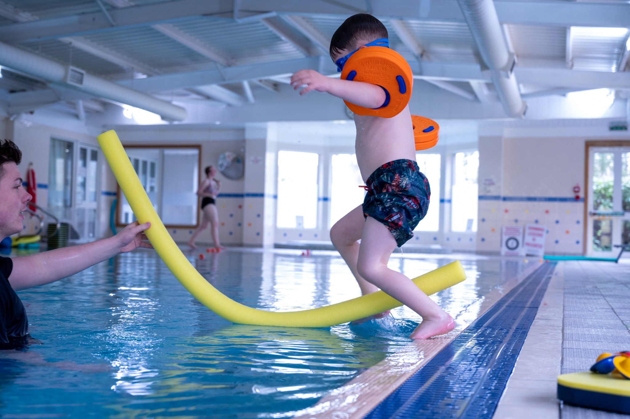 A swimmer jumps into the pool towards an instructor holding a pool noodle