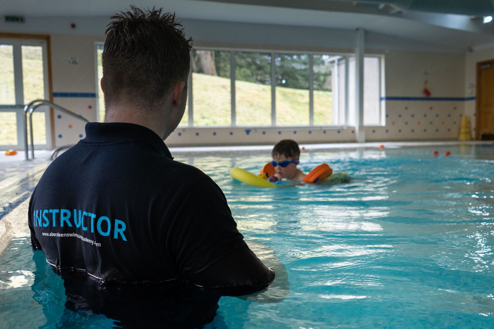 An instructor in the water watches a swimmer practise with a pool noodle