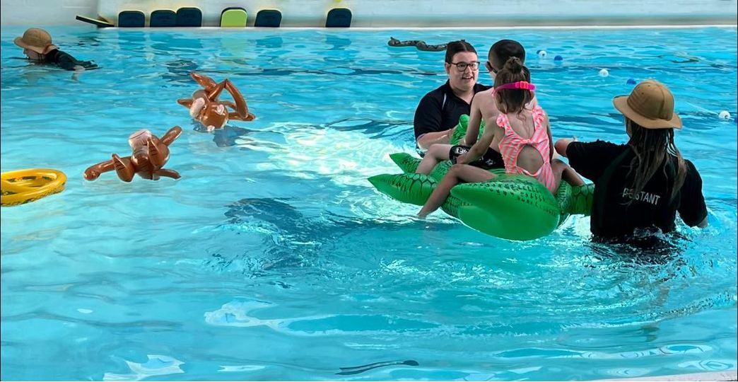 Staff supporting a child on a green float during a themed swimming lesson